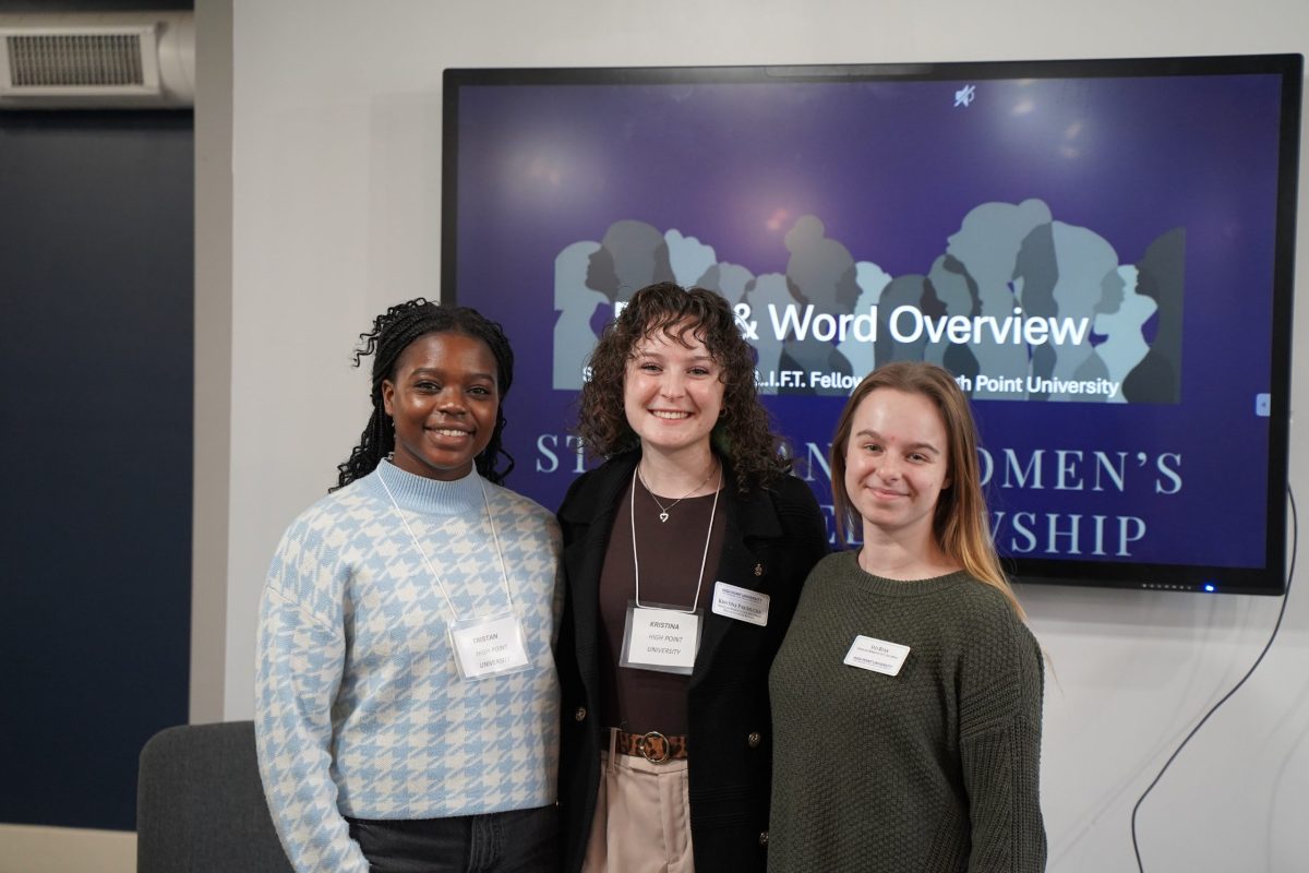 Three women stand indoors in front of a presentation screen displaying “Word Overview” at an event, all wearing name tags and smiling at the camera.