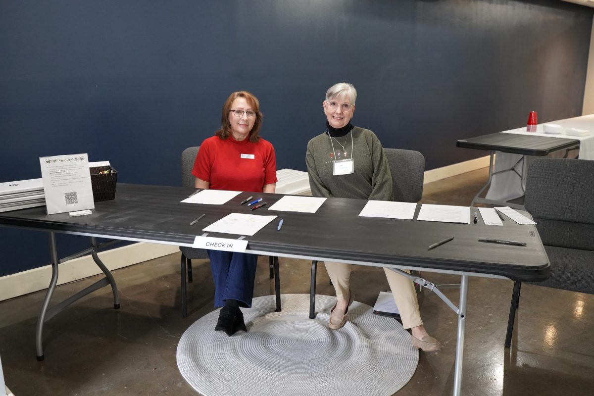 Two women sit behind a check-in table with papers and pens in a room with a blue wall. A sign reads “Check-In.”.