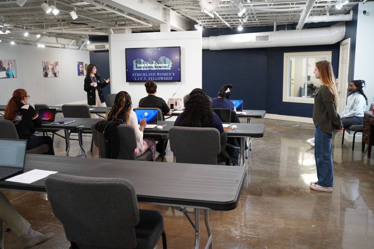 A group of people sit at tables facing a presenter speaking in front of a screen that displays “STRICKLAND WOMEN’S L.I.F.T. FELLOWSHIP.”.