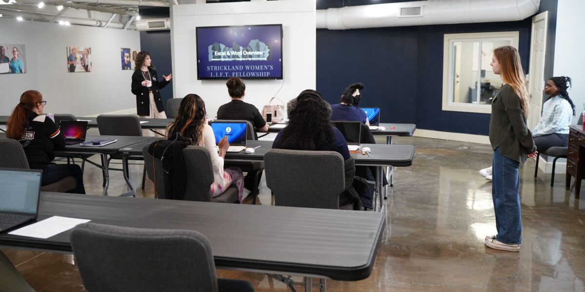 A group of people attend a presentation in a modern classroom. A speaker stands at the front near a screen displaying “Strickland Women’s L.E.A.P. Fellowship.”.