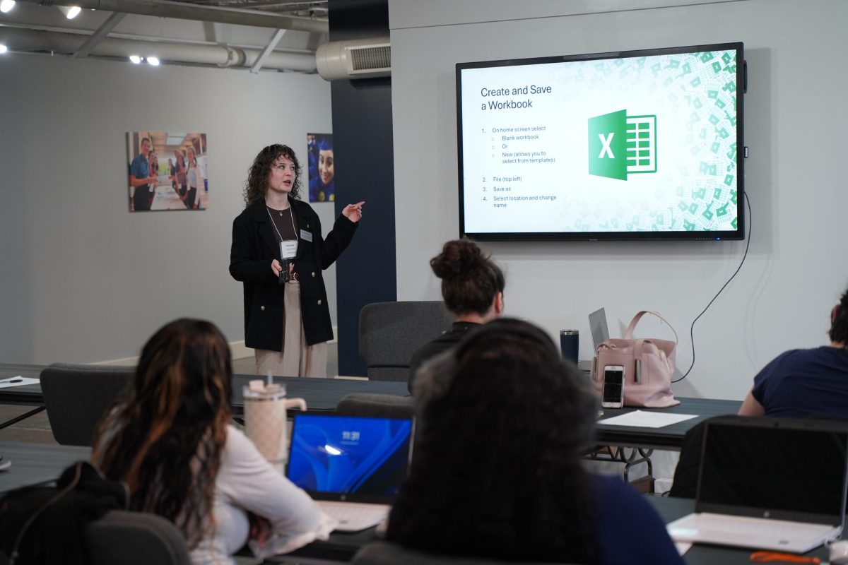 A woman presents a Microsoft Excel tutorial to a seated audience in a classroom, with a slide titled “Create and Save a Workbook” displayed on a large screen.