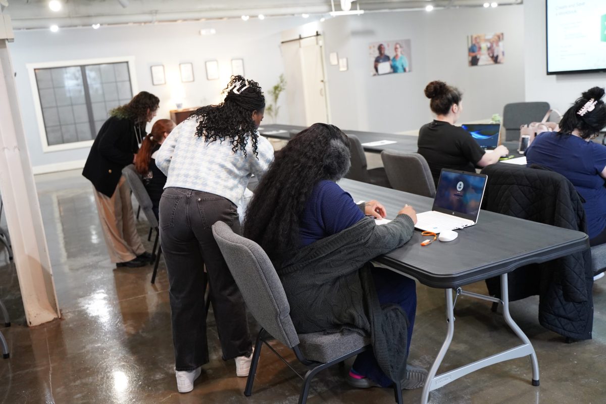 Several people are seated at tables in a classroom setting, some working on laptops, while others are engaged in discussion or assisting each other.