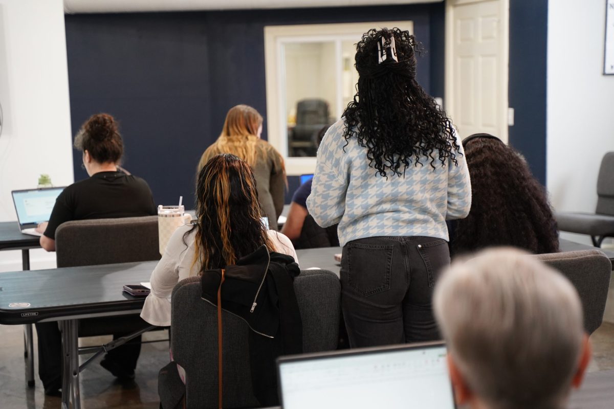 Several people, mostly seated, work on laptops in a classroom or office setting. One person stands near the center. All are viewed from behind.