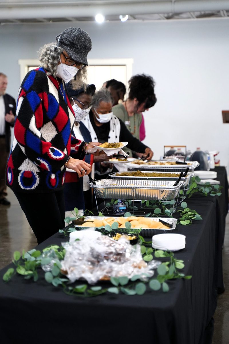 People wearing masks serve themselves food from a buffet table covered with various dishes and greenery in a well-lit indoor setting.