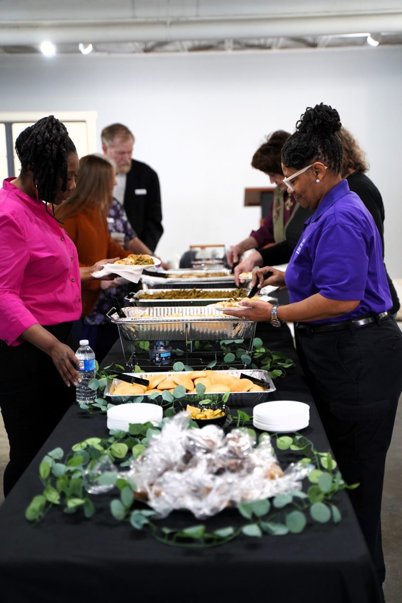 People serve themselves food from a buffet table covered with trays of dishes, plates, and bread, with greenery as decoration.