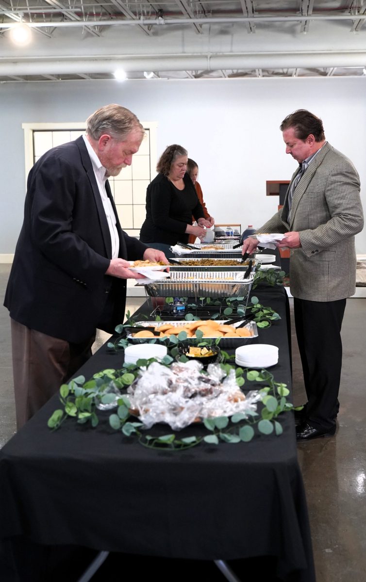 Two men serve themselves food from a buffet table covered with a black tablecloth and decorated with greenery, while two women stand behind the table.