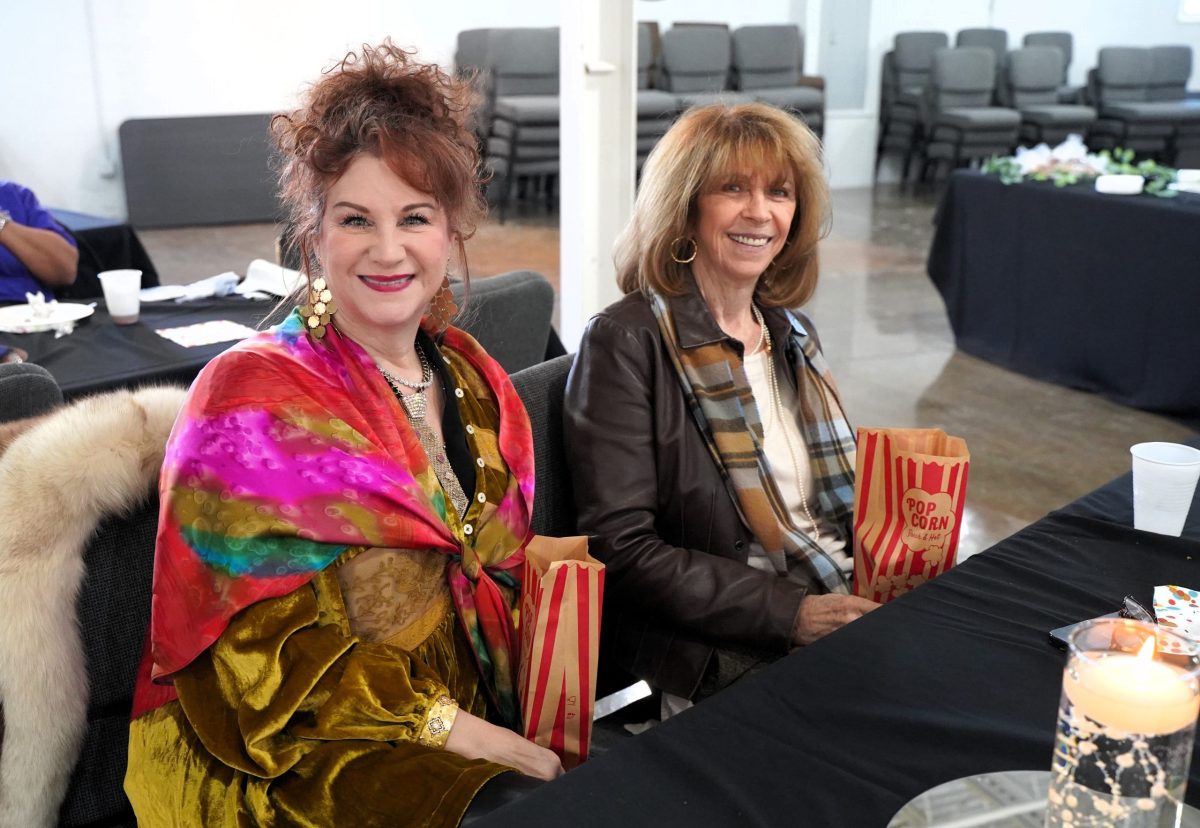 Two women sit at a table indoors, smiling and holding striped popcorn bags. There are empty chairs and tables with snacks in the background.
