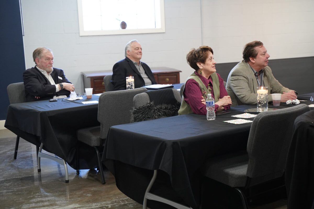 Four adults sit at tables covered in black cloth, facing forward in a well-lit room, appearing to listen attentively.