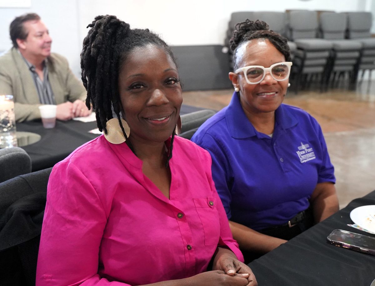 Two women sit at a table, one in a bright pink top and the other in a blue polo shirt. A man is seated in the background. They are indoors, possibly at an event or meeting.