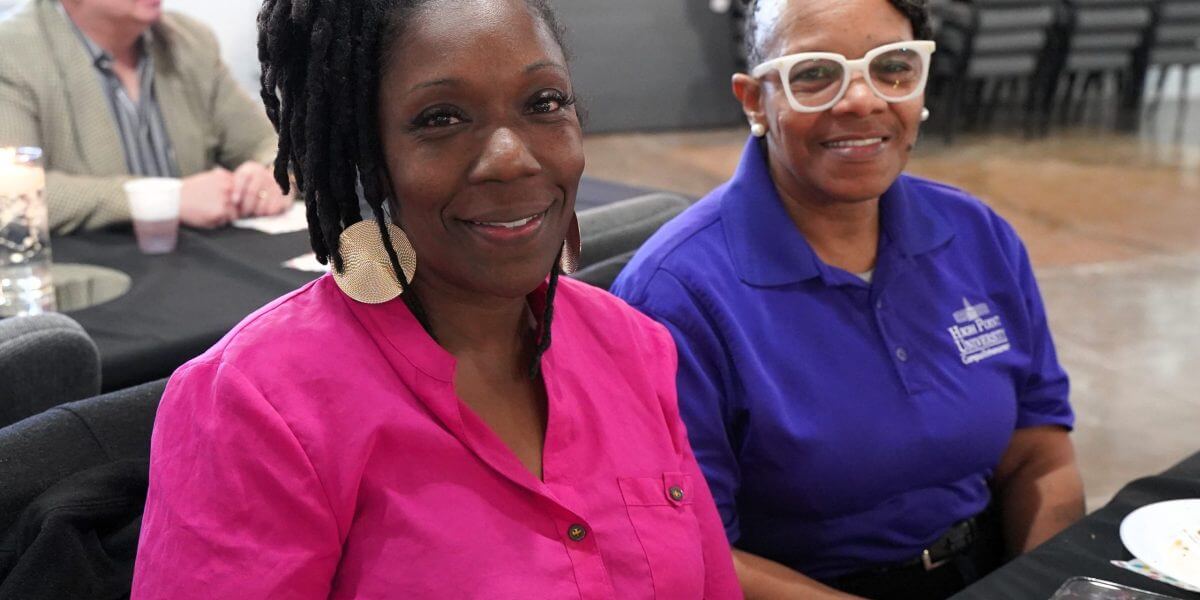 Two women sit side by side at a table, one wearing a bright pink top and the other in a blue polo shirt, with a man seated in the background.