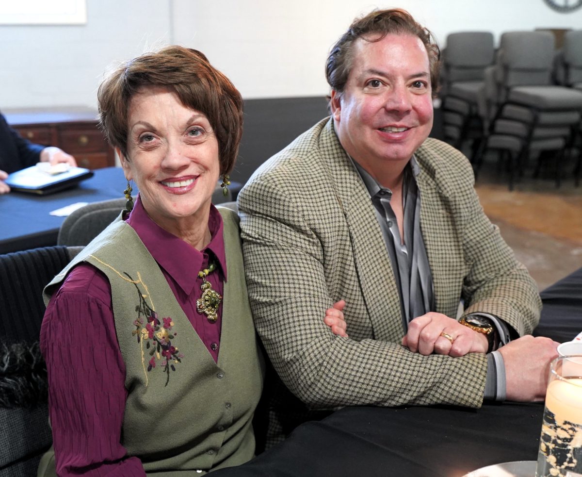 A woman and a man sit next to each other at a table, smiling at the camera in a well-lit indoor setting with chairs in the background.