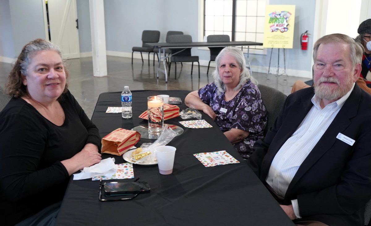 Three adults sit at a black table with snacks, drinks, and napkins in a bright room. A sign and empty chairs are visible in the background.