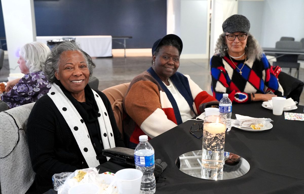 Three women sit at a black table with drinks and snacks, smiling at the camera in a bright indoor space.