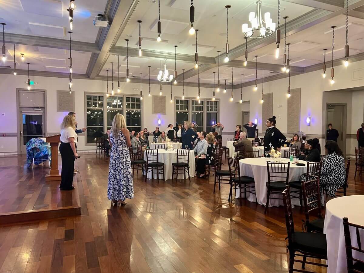 People are gathered in a spacious, well-lit banquet hall with round tables and wooden floors, some seated and some standing, during an indoor social event or reception.