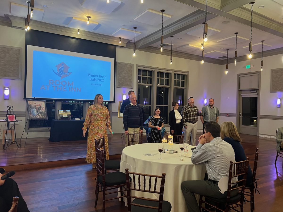 Five people stand at the front of a banquet room, speaking to seated guests. A large screen behind them displays “Room at the Inn” and “Winter Rose Gala 2023.”.