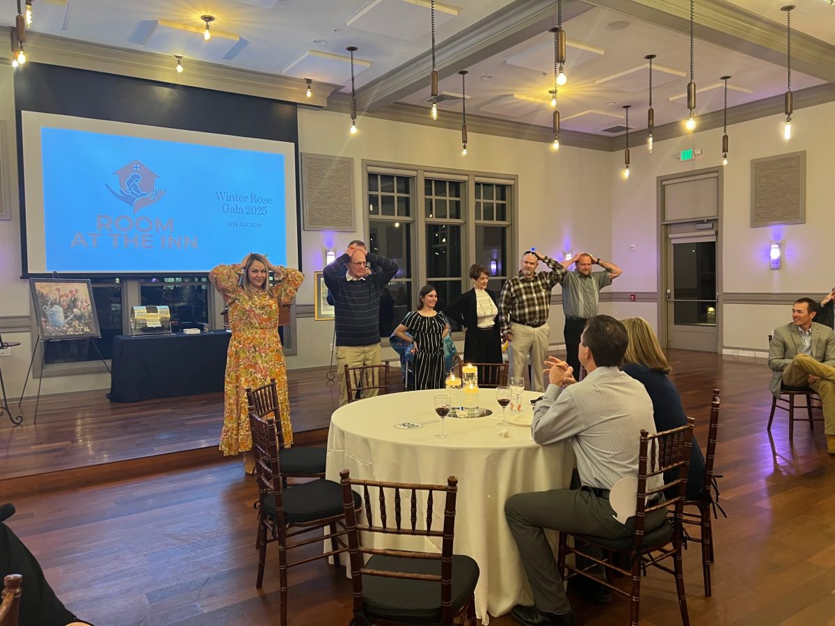Five people stand on stage miming moose antlers as others watch from tables at an indoor event; a screen reads “Room at the Inn Winter Soiree Gala 2023.”.