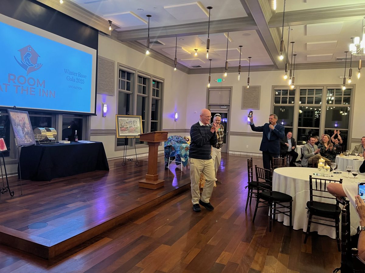 A man walks across a banquet room as another man speaks to an audience. Art is displayed on tables, and a large screen reads “Room at the Inn, Winter Rose Gala 2023.”.