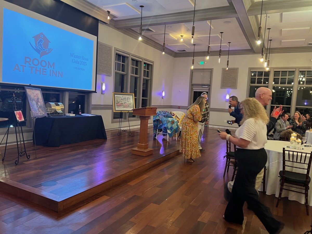 A woman bows on stage at a Room at the Inn gala, while people watch and take photos. A screen, artwork, and tables are visible in the event hall.