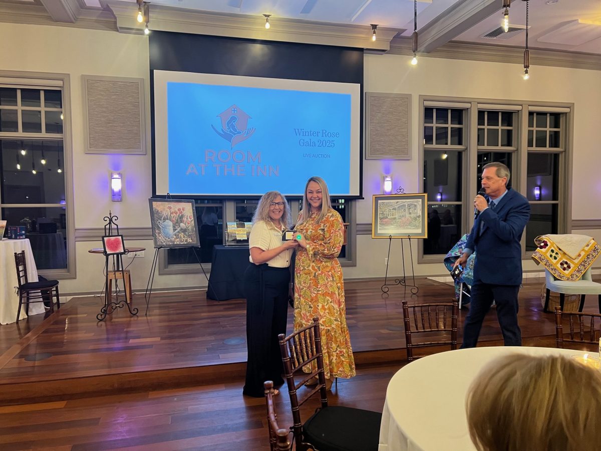 Two women stand together holding an award at a gala event, while a man with a microphone speaks on stage. A presentation screen behind them displays “Room at the Inn Winter Rose Gala 2023.”.