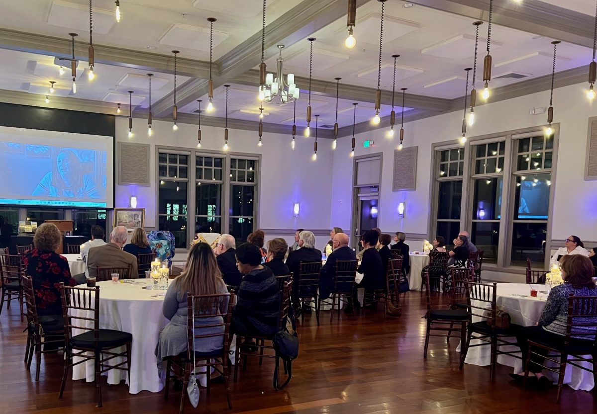 People seated at round tables in a well-lit banquet hall watch a video presentation projected onto a screen at the front of the room.
