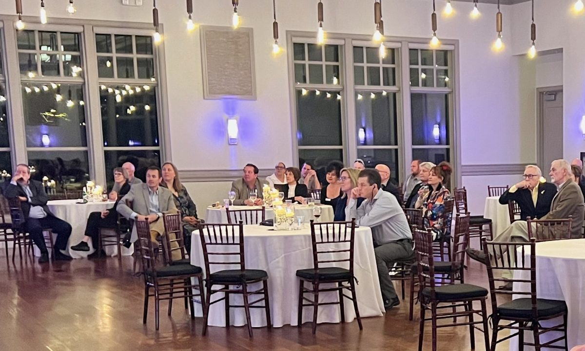 People seated at round tables in a well-lit event space, attentively watching something off-camera. Hanging lights and large windows are visible in the background.