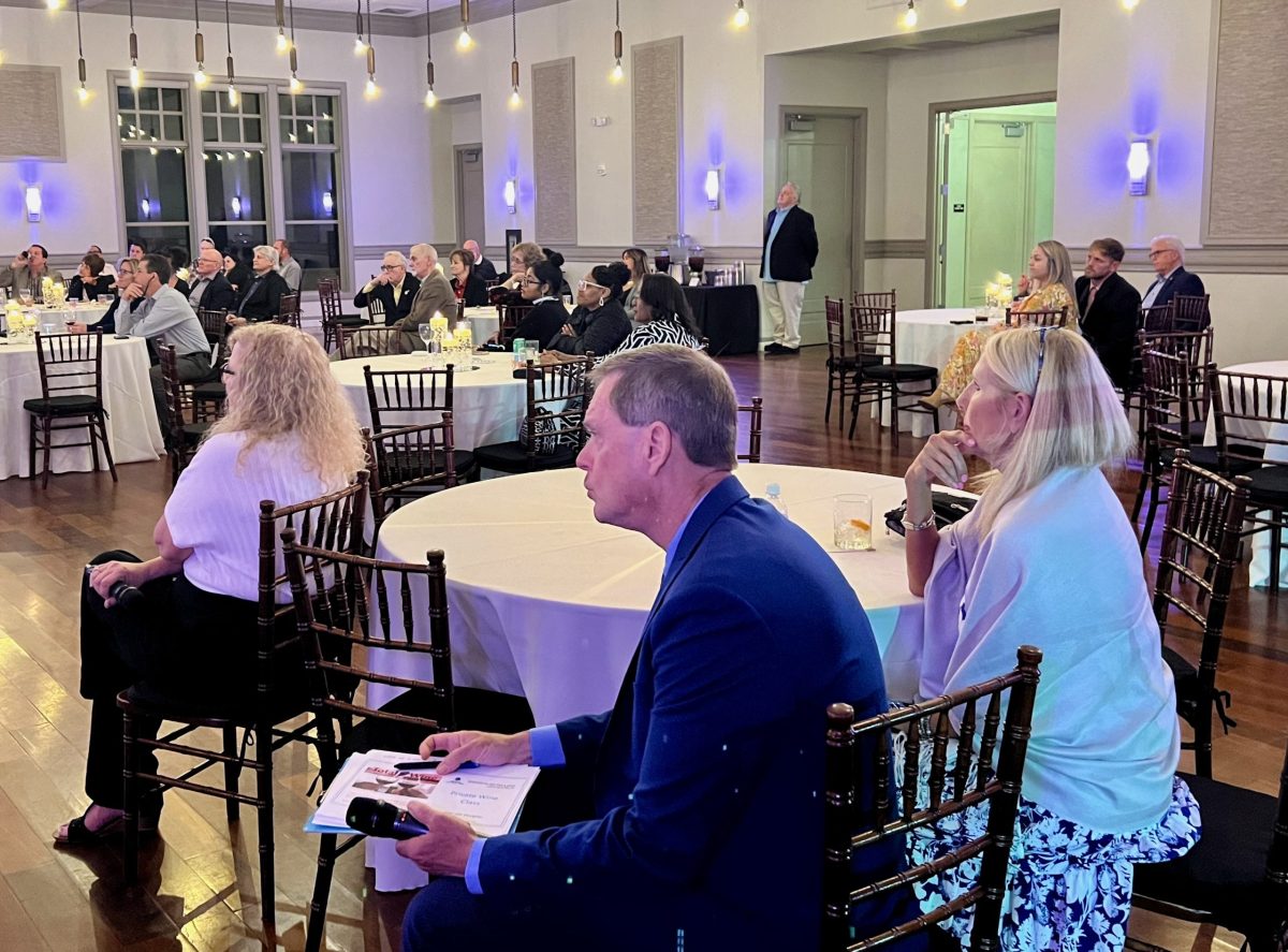 People seated at round tables in a decorated event hall, attentively watching a presentation or speaker. The room has wooden floors, hanging lights, and formal attire.