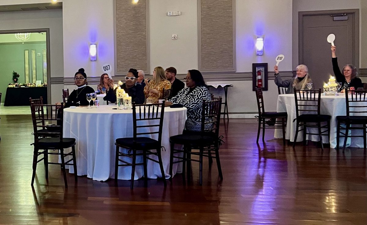 People sit at round tables in a banquet hall, some holding numbered paddles, suggesting a live auction or bidding event.