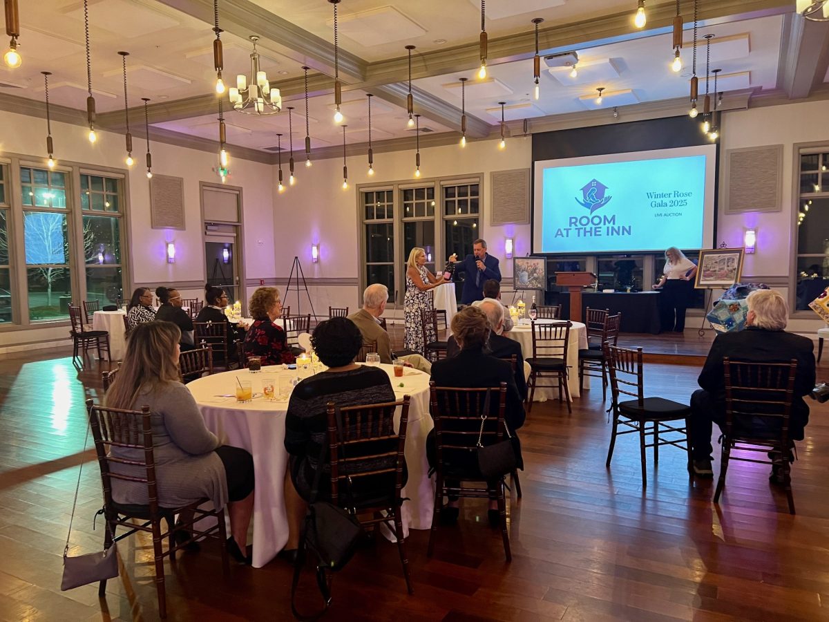 People seated at round tables watch a speaker at a podium in a well-lit banquet hall with a “Room at the Inn” presentation displayed on a large screen.