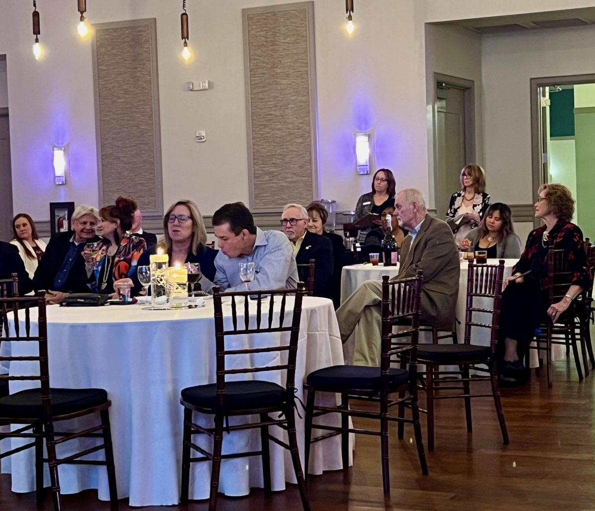 People sit at round tables in a banquet hall, listening attentively. The room has wooden chairs, white tablecloths, and wall sconces for lighting.