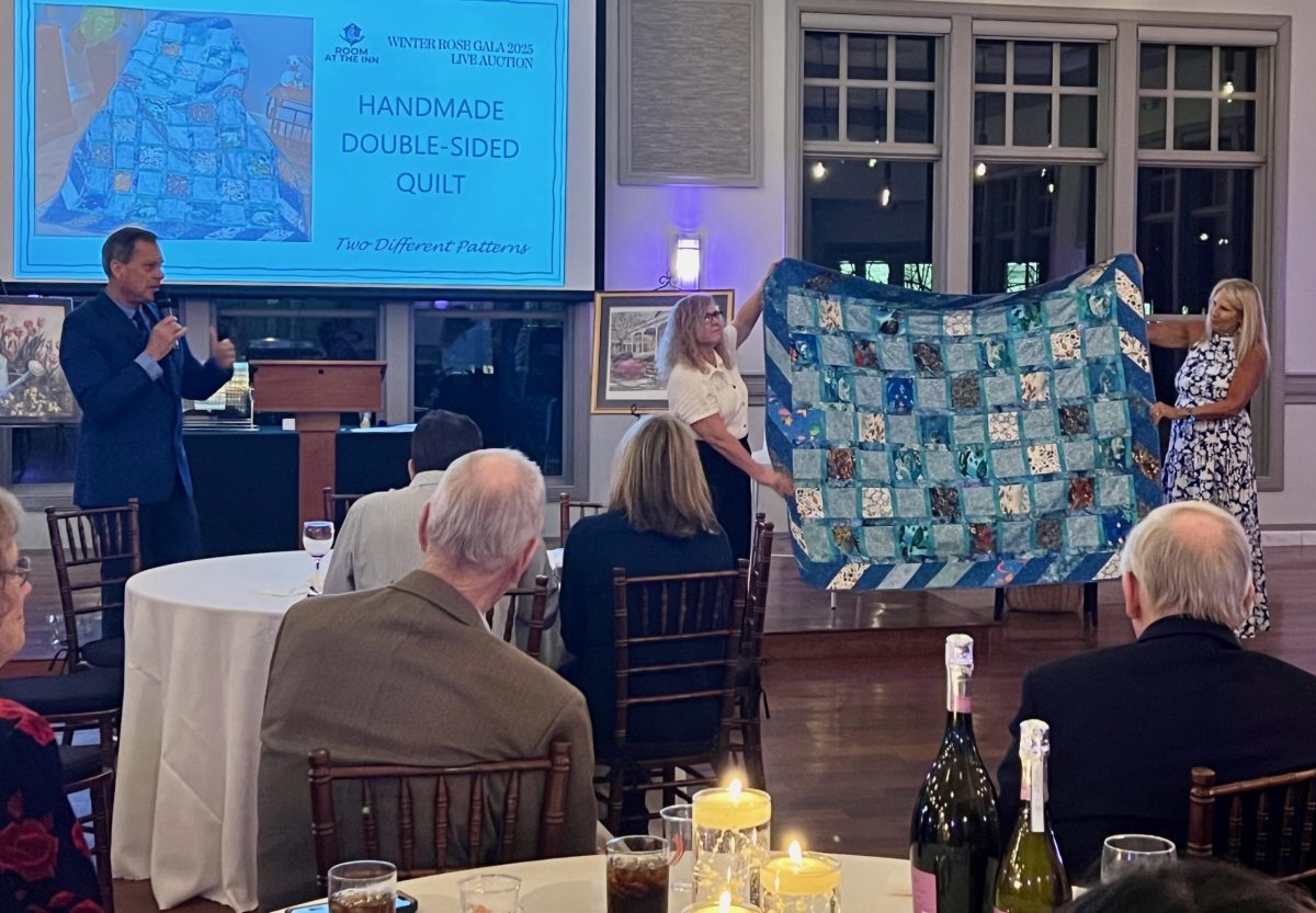 Two women display a handmade double-sided quilt at an auction, as a man speaks at a podium. Guests are seated at tables with candles, wine bottles, and glasses in the foreground.