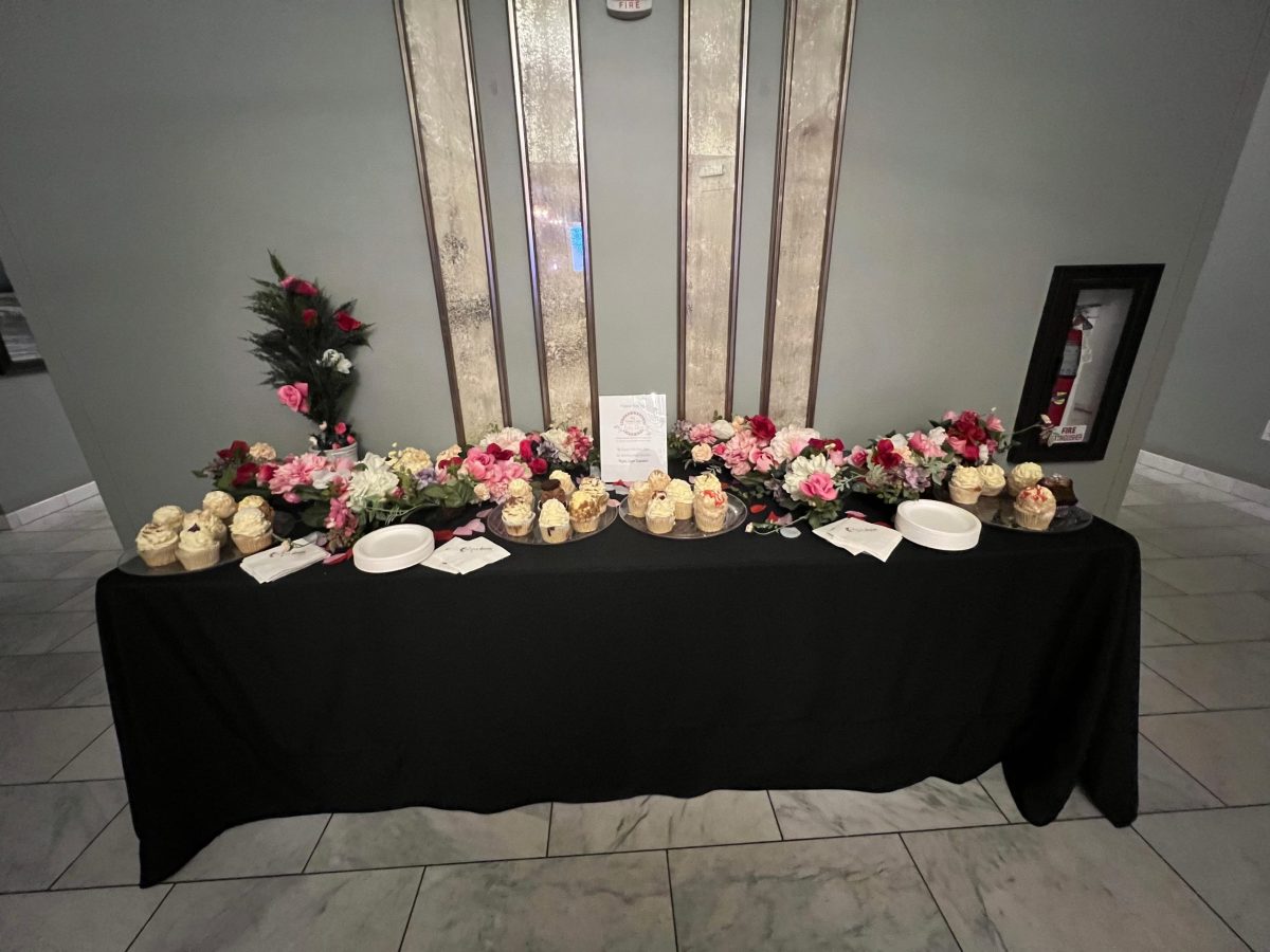 A table with a black tablecloth displays cupcakes, plates, napkins, floral arrangements, and a sign against a gray wall.