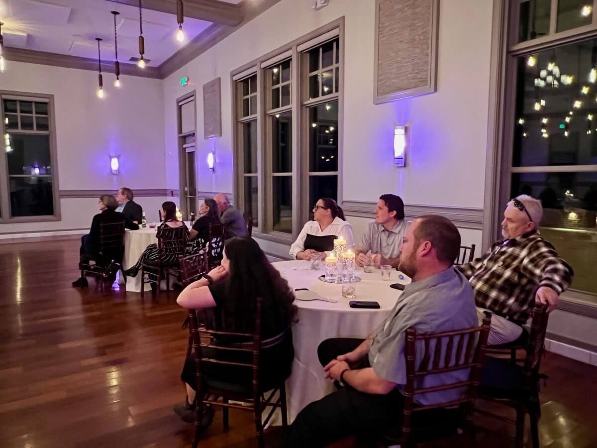 People sit around tables in a softly lit banquet room, watching something off-frame. The room has large windows, wood floors, and several pendant lights hanging from the ceiling.