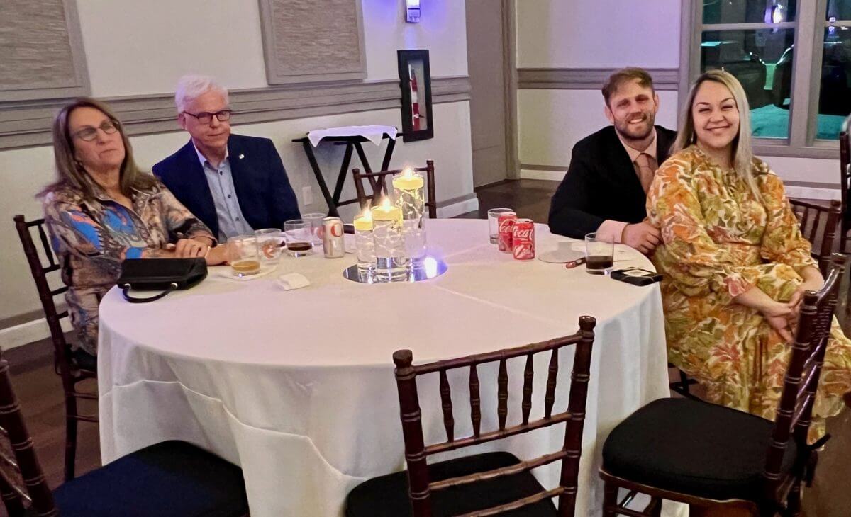 Four people sit at a round table with drinks and candles in a decorated indoor event space. Empty chairs surround the table.