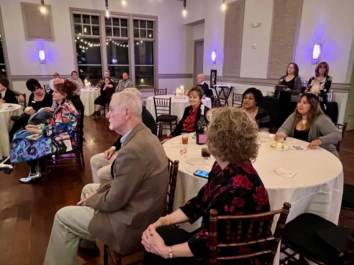 People seated around round tables at an indoor event, attentively watching something off-camera; drinks and plates are visible on the tables.