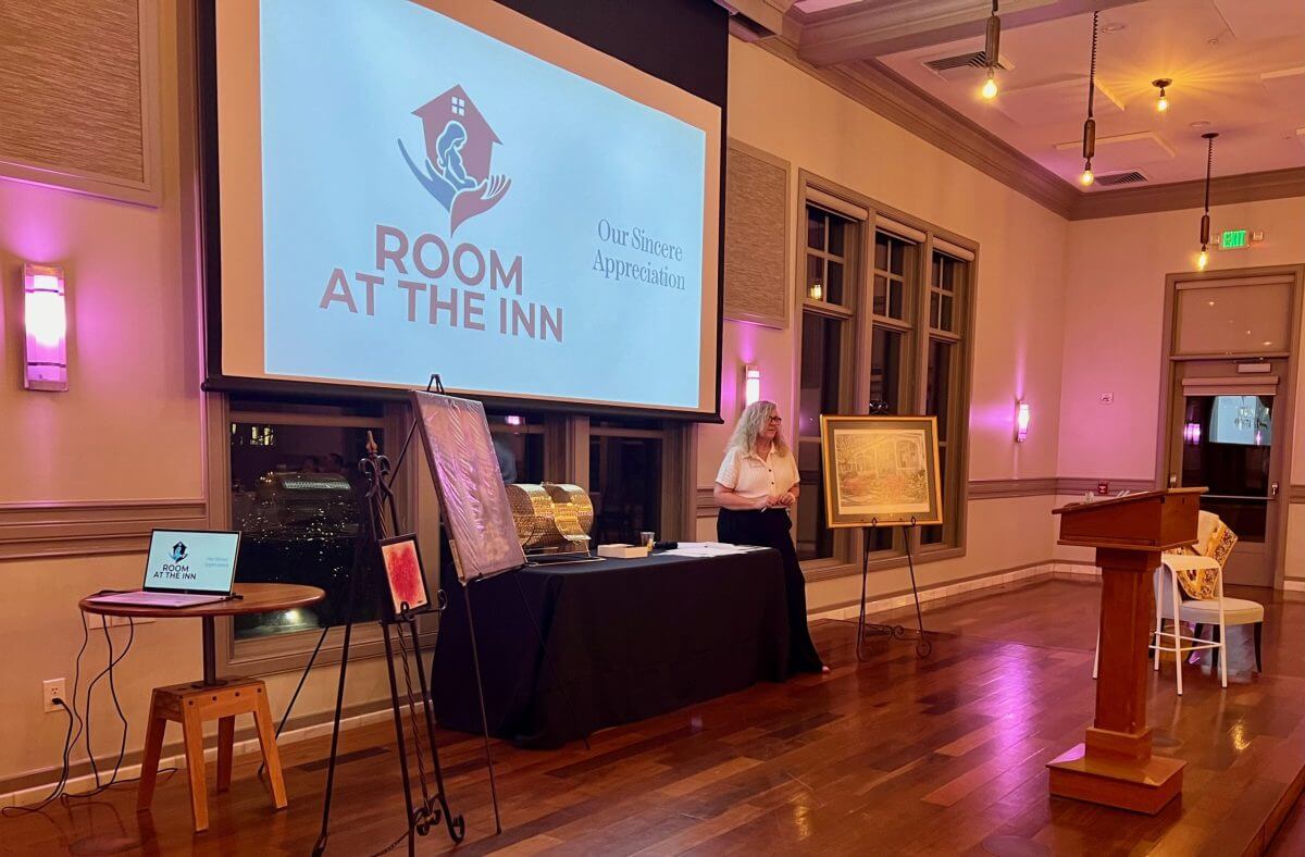 A woman stands next to a table and artwork in a room with a large screen displaying the “Room at the Inn” logo and message of appreciation.