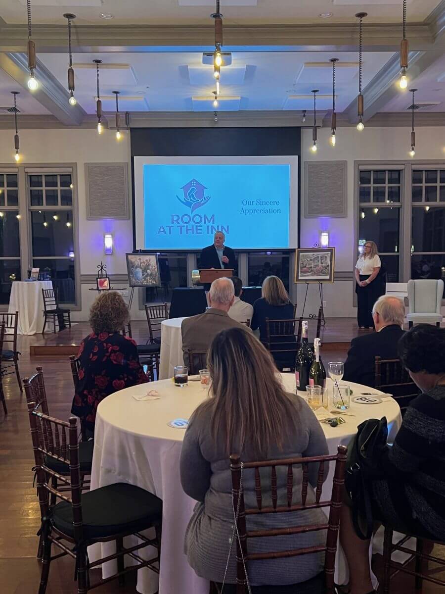 A speaker addresses seated guests at a formal event with a “Room at the Inn” presentation displayed on a screen at the front of the room.