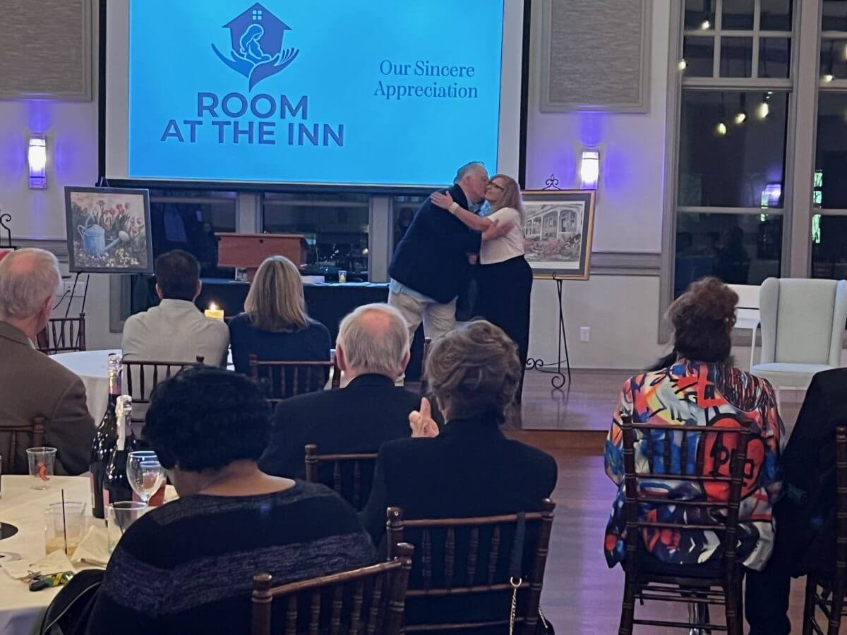 A man and woman hug onstage at a Room at the Inn event, with seated guests watching and framed artwork displayed on easels beside them.