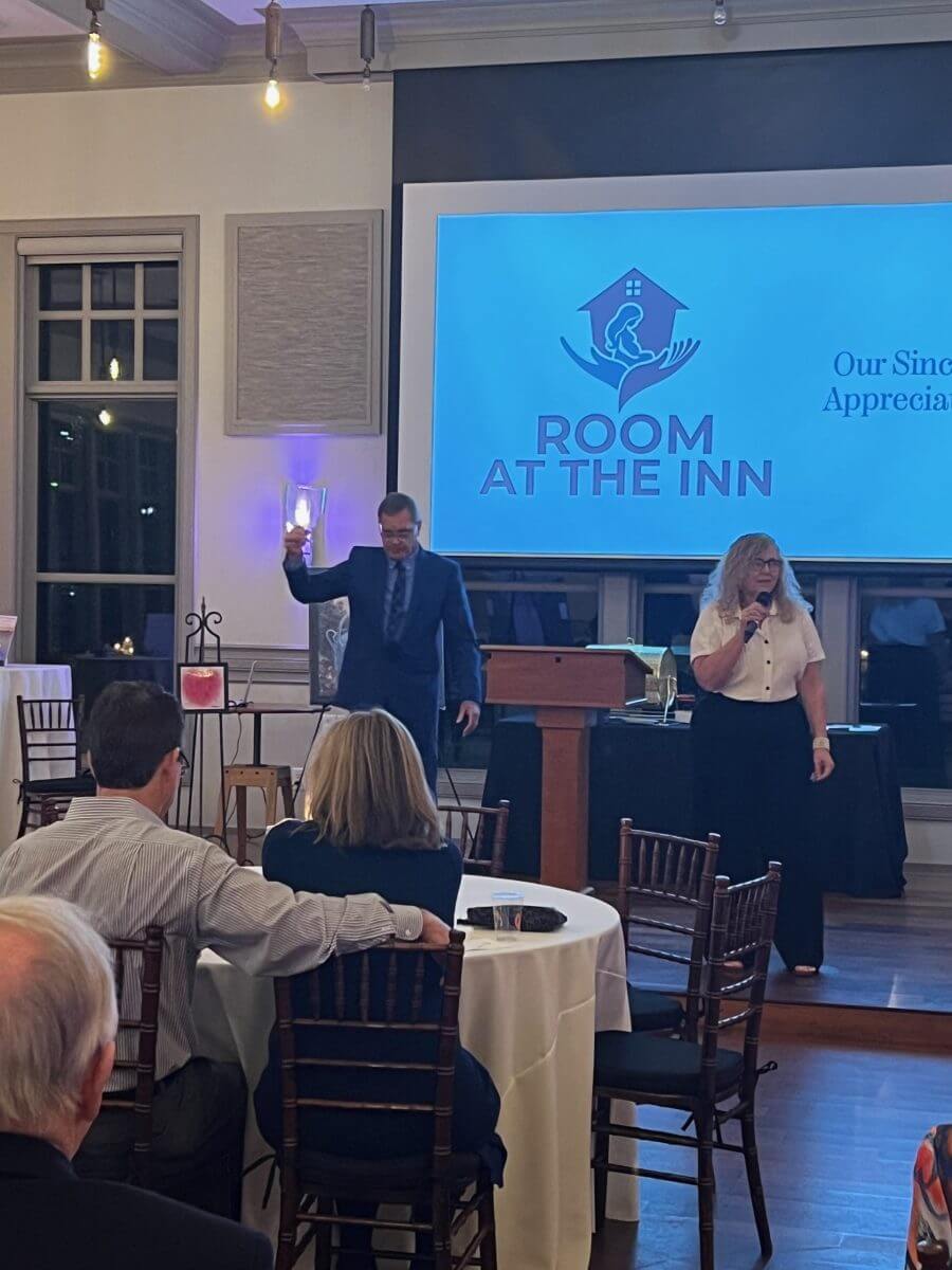 A man and woman stand on stage at a “Room at the Inn” event; the man holds up an object while the woman speaks into a microphone. Audience members are seated at round tables.