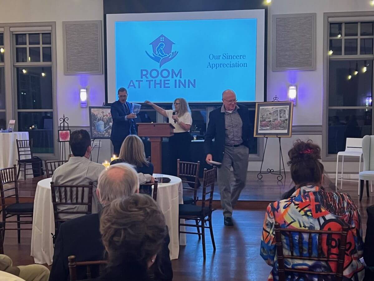 A woman speaks at a podium with two men nearby, one holding a painting, at a “Room at the Inn” event in a banquet hall with attendees seated at round tables.