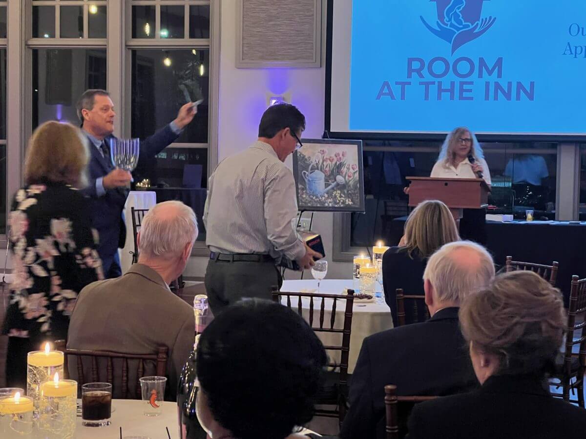 A man holds up a menorah while another holds a box at a formal event. A woman speaks at a podium near a “Room at the Inn” sign. Guests sit at candlelit tables.