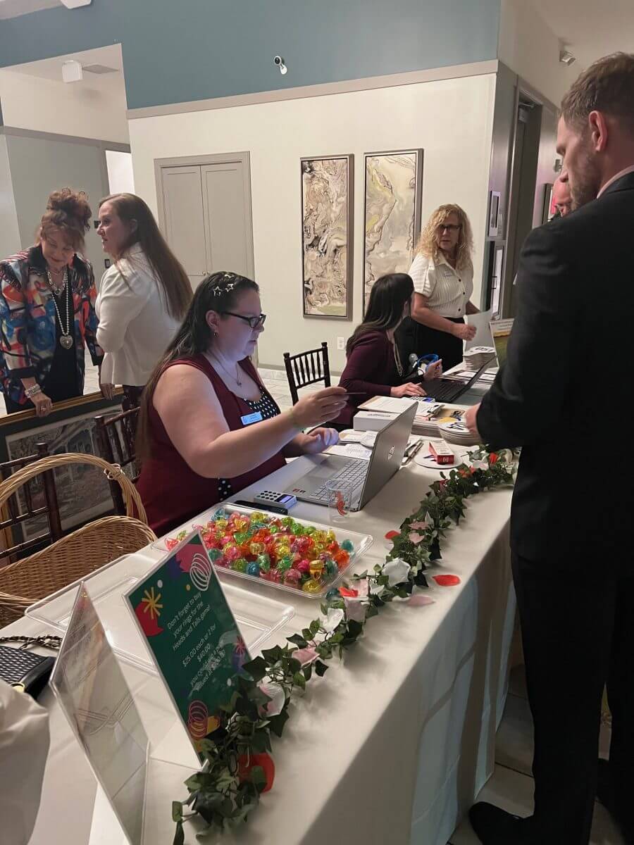 People interact at a registration table with laptops and candy dishes in a decorated indoor space. Artworks hang on the wall in the background.