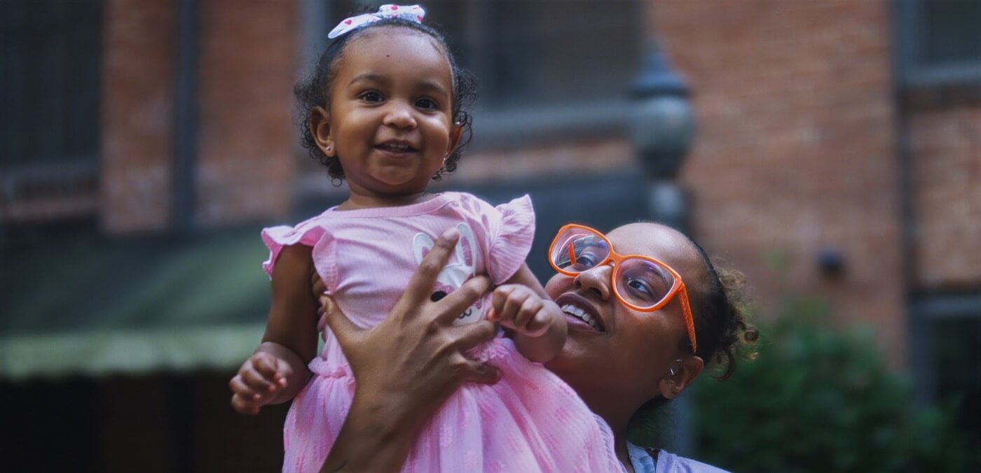 An adult wearing orange glasses lifts a smiling toddler in a pink dress outdoors, with a brick building in the background.