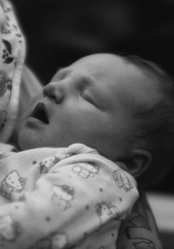 A baby in patterned pajamas sleeps with mouth slightly open, resting against an adult holding a floral-patterned cloth in the comforting environment of a maternity shelter in Greensboro, NC.